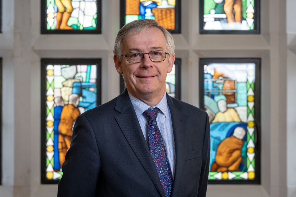 Emeritus Professor Chris Linton, wearing a suit, is stood in front of a stained glass window