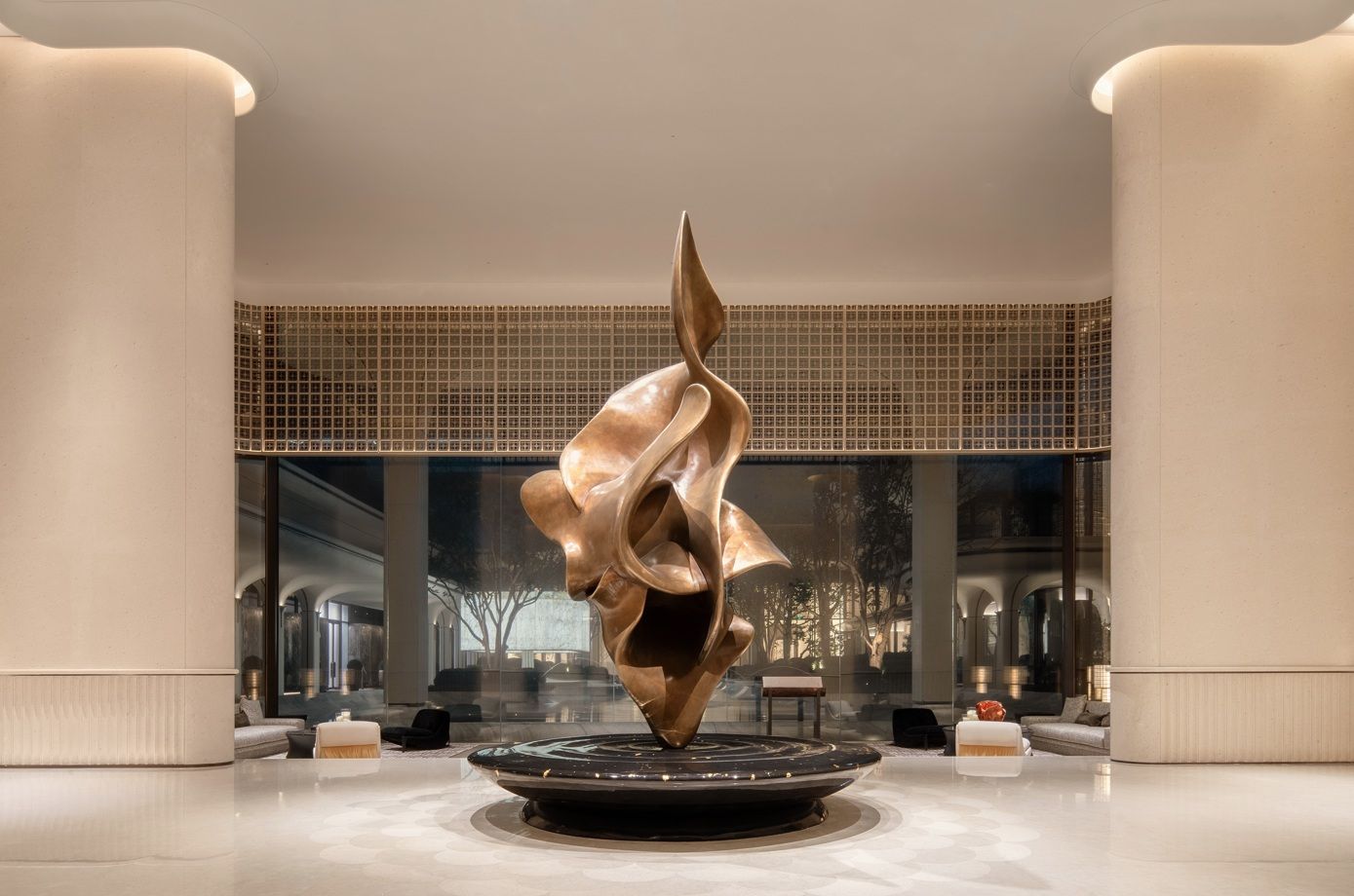 A large abstract metallic sculpture stands on a circular base inside a grand building with cream walls, and a marble floor.