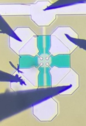 Close-up of a microchip with a symmetrical cross-shaped pattern, surrounded by several probing needles used to measure its electrical behaviour.