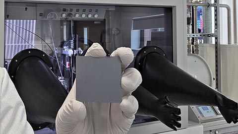 A hand wearing white gloves holding up a piece of solar panel in a research laboratory