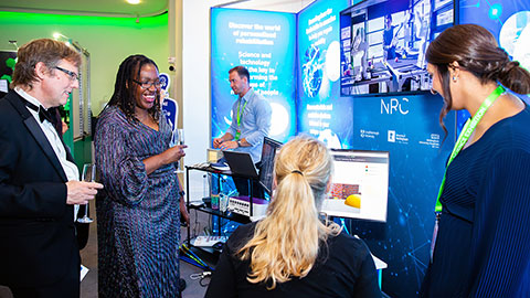 Two guests holding drinks talking to a person sitting at a computer and a second person standing against a backdrop of the exhibition