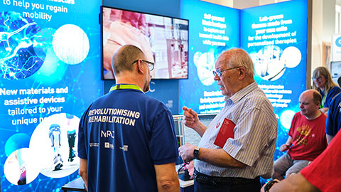 A guest talking to a person wearing a t-shirt with the words 'Revolutionising Rehabilitation' printed on the back with display stands and a digital screen in the background