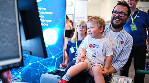 A man with a child on his knee sitting on an interactive seat laughing while watching a digital screen