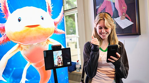 A girl with her thumb up holding a mobile phone, alongside a tablet on a tripod and a banner with a photo of an axolotl in the background