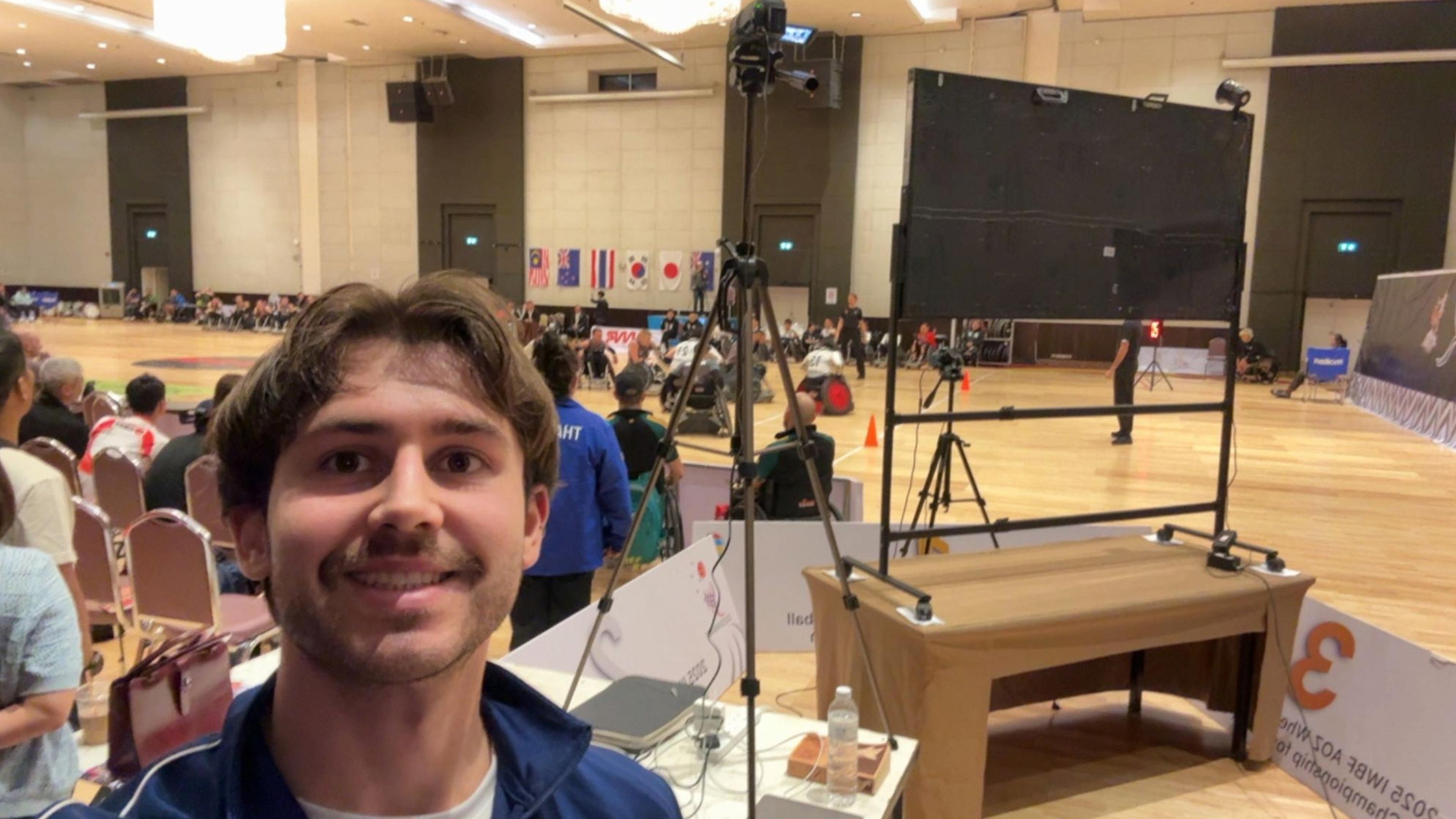 A photo of Owen Tolfrey stood in front of a wheelchair rugby court whilst a game is taking place