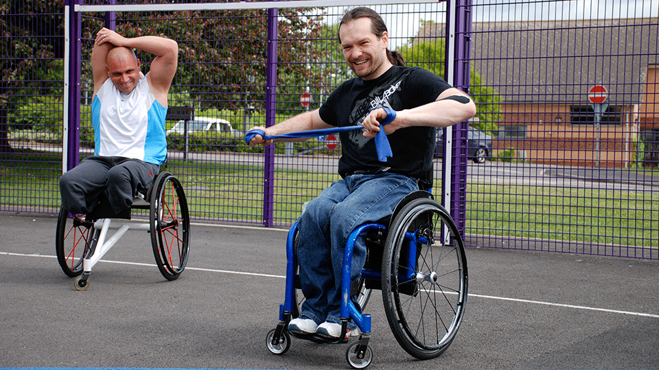 Two male wheelchair users enjoying some physical activity using resistance bands outside.