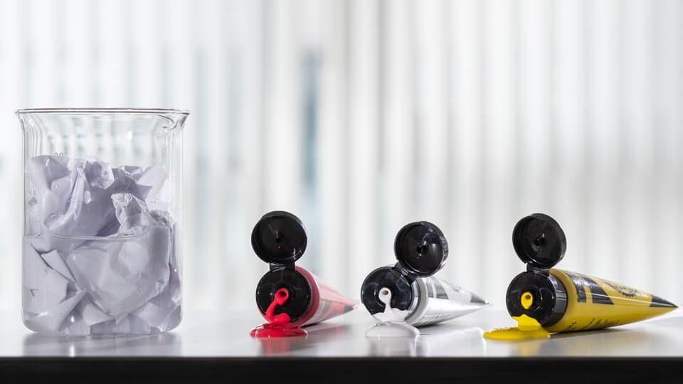 A beaker of paper sludge beside three tubes of acrylic paint