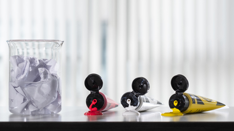 A beaker of paper waste beside three tubes of acrylic paint