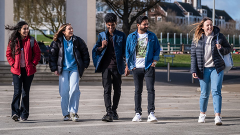 A group of five students walking towards the camera on campus with buildings in the background