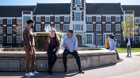 Three postgraduate students sitting and standing talking in front of the fountain and Hazlerigg Building
