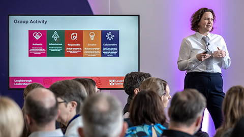 A presenter standing in front of an audience of people alongside a large display screen