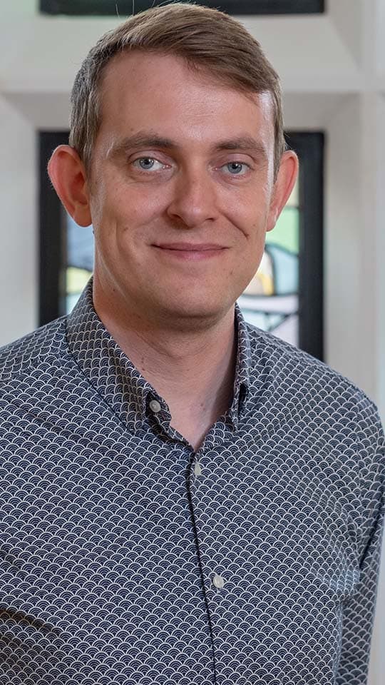 A portrait photo of Stuart King standing in front of a stained glass window