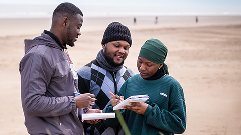 A group of three students talking and making notes on a beach