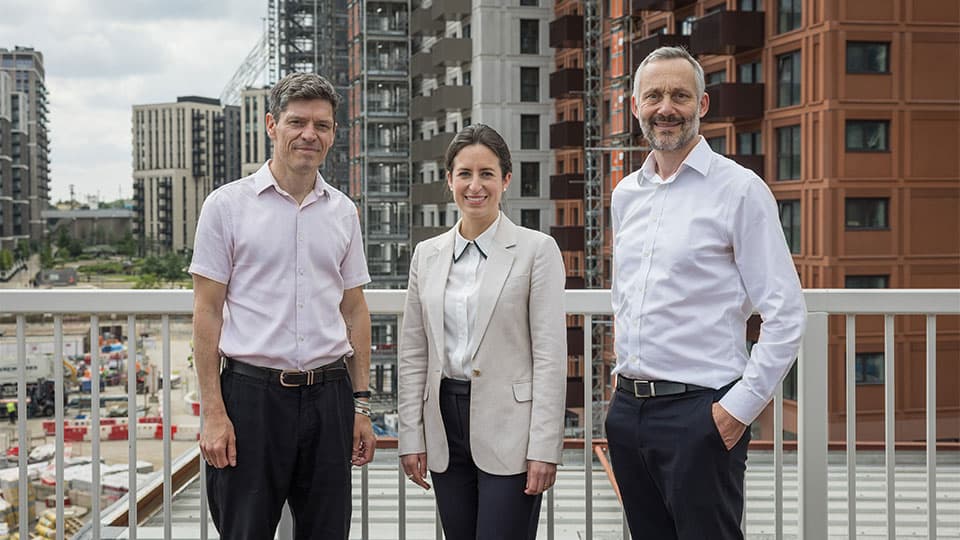 Chris is standing with a lady and a man in front of tall buildings outside. He is wearing a short sleeved shirt and dark trousers.
