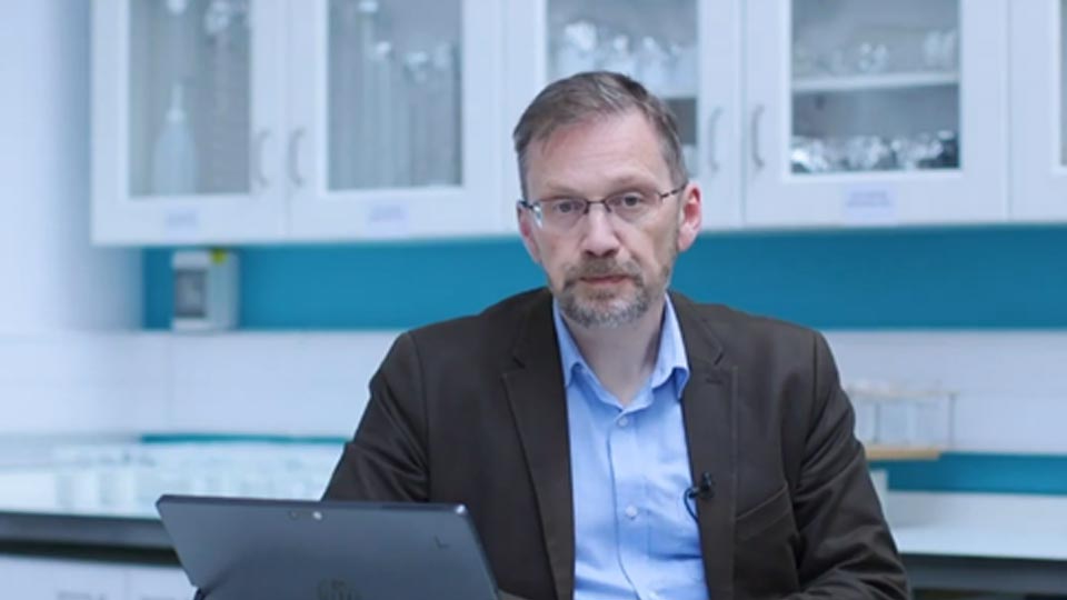 Dr Hopkins sitting with dark suit and light blue shirt and glasses in front of laptop facing camera