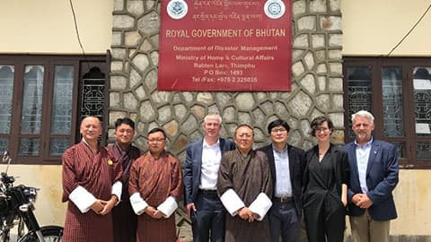 A group of people stand for a photo outside a building with a sign which reads: Royal Government of Bhutan, Department of Disaster Management, Ministry of Home and Cultural Affairs.