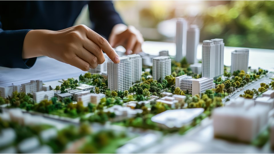 A female is arranging a model of a small building on a horizontal board on a table covered with urbanised buildings and greenery inside a studio.