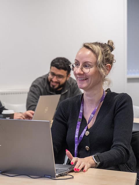 A professional education student working at a laptop, smiling at something off camera.