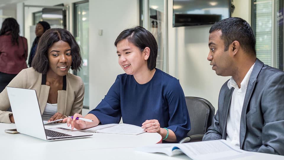 A group of three students sitting at a table looking at a laptop screen.