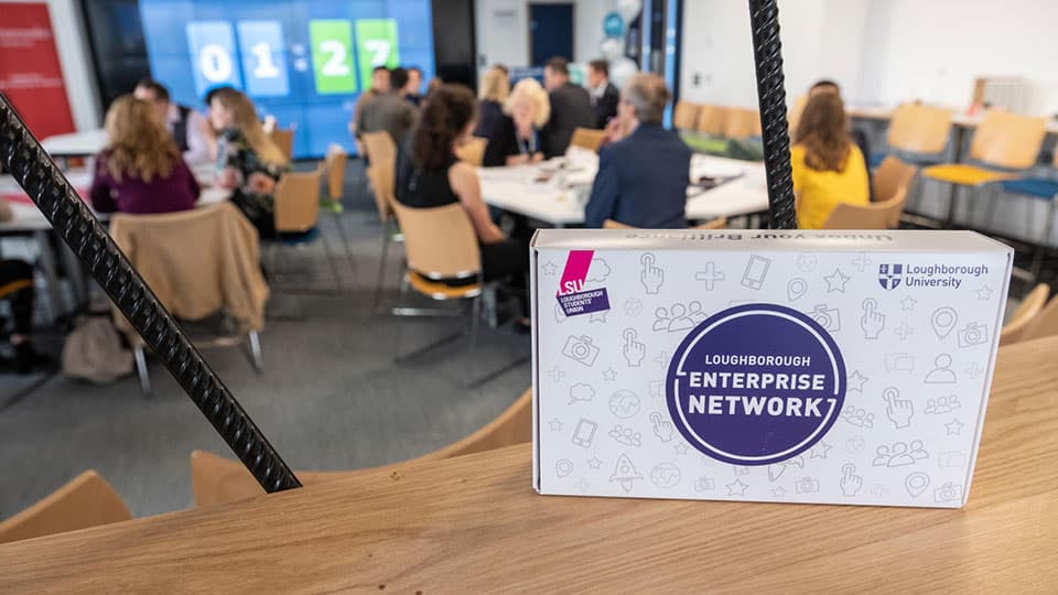 A white box with the Loughborough Enterprise Network logo on a shelf, with people sitting and chatting in the background.