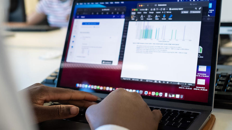 A person sitting at a desk using a laptop with graphs and charts on screen