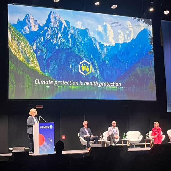 Professors Tom Jackson and Ian Hodgkinson on stage at the HIMSS 2024 conference. There is a large screen behind them with a photo of a mountain and text that reads 'Climate protection is health protection'.