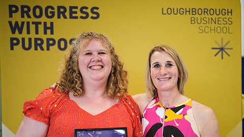 Malika Lawrance holding their award and certificate with Jan Godsell. They are stood in front of a bright yellow banner.