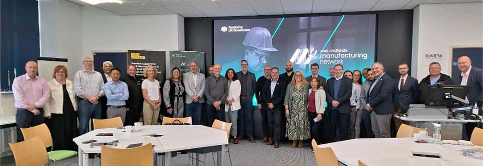 Delegates from the Leicestershire Manufacturing Network launch event standing in a row. Behind them is a large screen with an East Midlands Manufacturing Network slide.