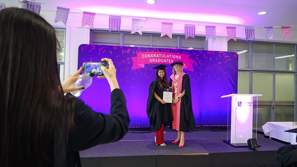 A graduate on stage holding a certificate from the Dean of Loughborough Business School. There is someone in the foreground taking a photo of them on a smartphone.