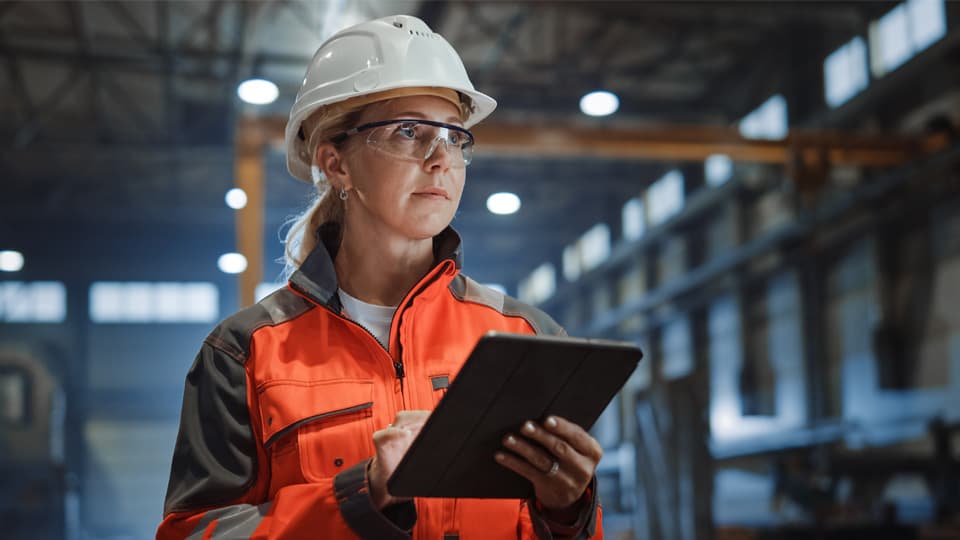 A worker in an industrial building, wearing a hard hat, hi vis clothing, and holding a tablet computer.