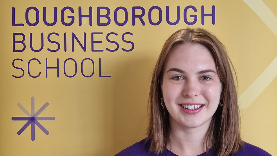 A person standing in front of a yellow backdrop with the text 'LOUGHBOROUGH BUSINESS SCHOOL' in purple.