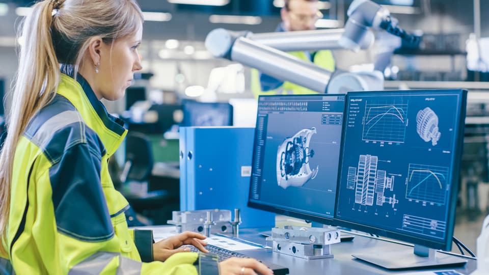 a woman standing at a desk looking at two computer screens