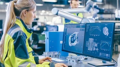 a woman standing at a desk looking at two computer screens