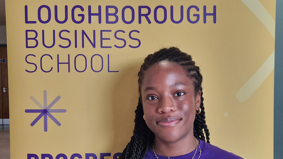 A person standing in front of a yellow backdrop with the text 'LOUGHBOROUGH BUSINESS SCHOOL' in purple.