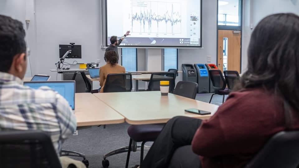People in a seminar, sat around tables watching a presenter at the front.