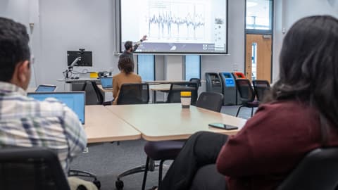 People in a seminar, sat around tables watching a presenter at the front.