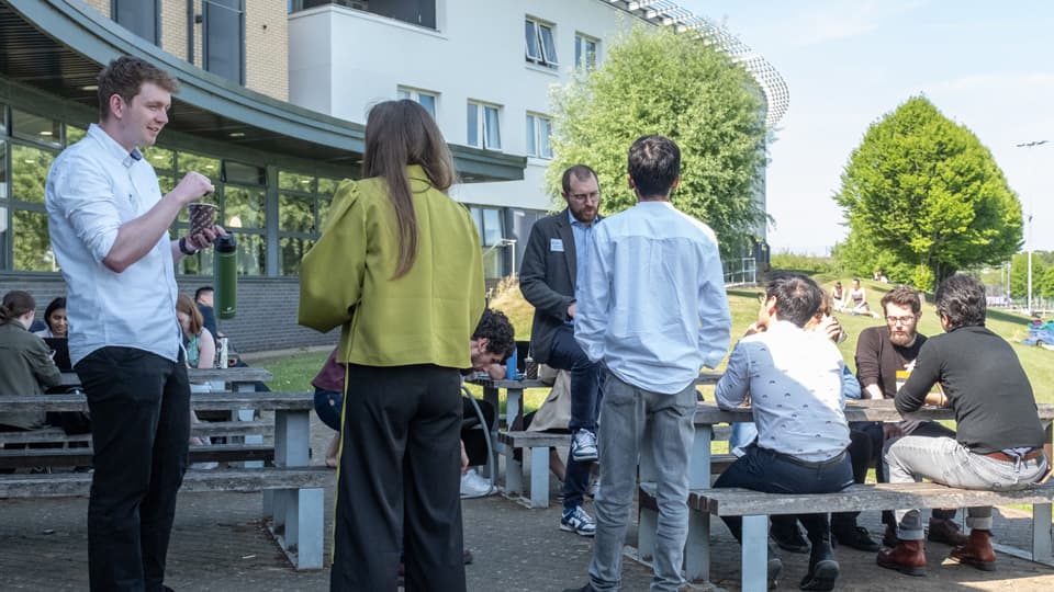 People gathered at an outdoor seating area on a sunny day, sitting, standing and chatting together.
