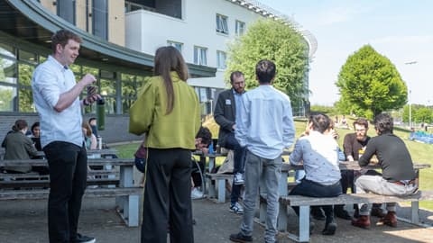 People gathered at an outdoor seating area on a sunny day, sitting, standing and chatting together.