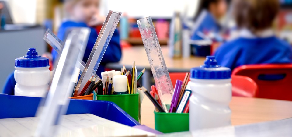 Classroom desks with containers of school supplies like rulers, pencils, and markers, with students in blue uniforms blurred in the background.