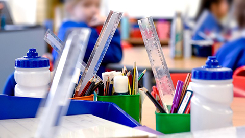 Classroom desks with containers of school supplies like rulers, pencils, and markers, with students in blue uniforms blurred in the background.