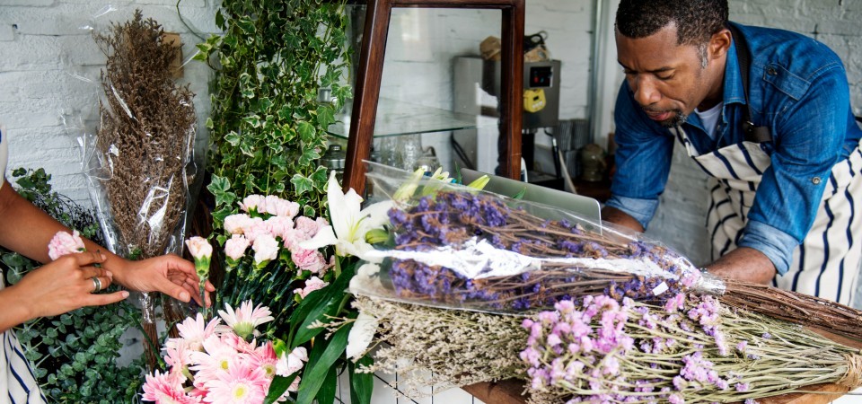 Two people arranging flowers in a florist shop with a laptop and various blooms on the table.
