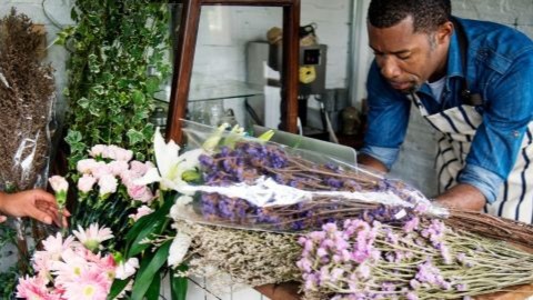 Two people arranging flowers in a florist shop with a laptop and various blooms on the table.