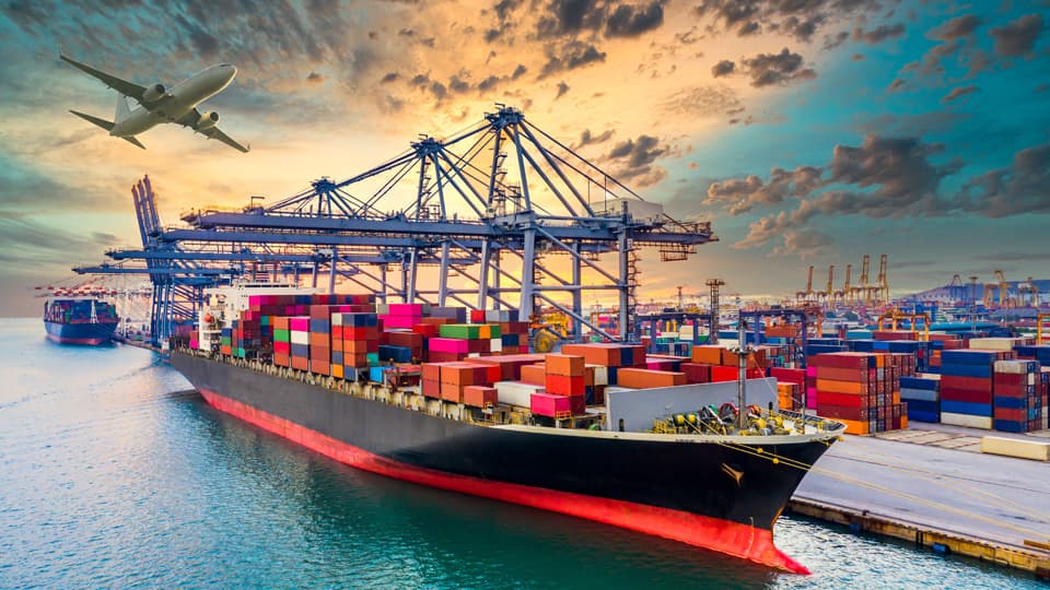 A cargo ship docked at a cargo port, with an aeroplane in flight in the background.
