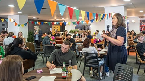University staff and students gathered in an open space. A staff member is giving a speech. The students are sitting around tables. The venue is decorated with colourful bunting.