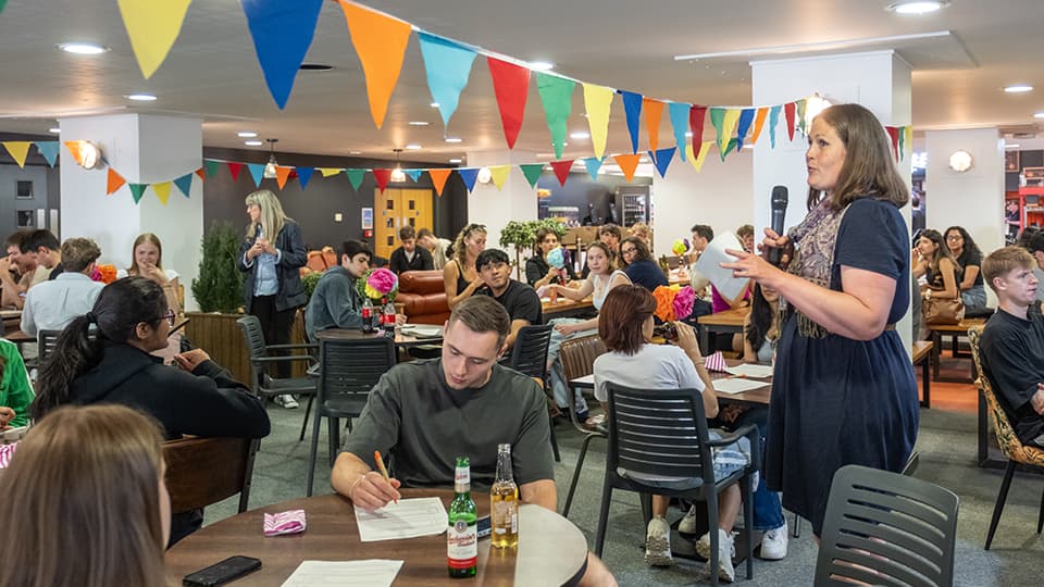 University staff and students gathered in an open space. A staff member is giving a speech. The students are sitting around tables. The venue is decorated with colourful bunting.