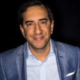 Luq Niazi wearing a navy blue jacket and light coloured shirt against a dark background