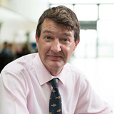 Neil Stansfield wearing a light coloured shirt and dark tie sitting a desk