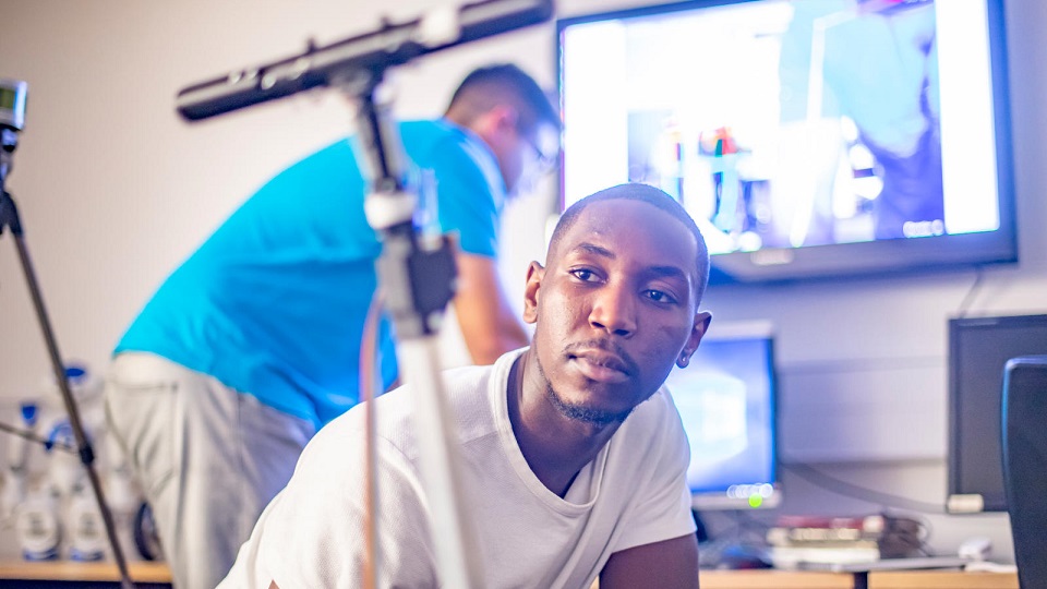 Male student with virtual reality equipment, with another male student blurred in the background stood at a computer.