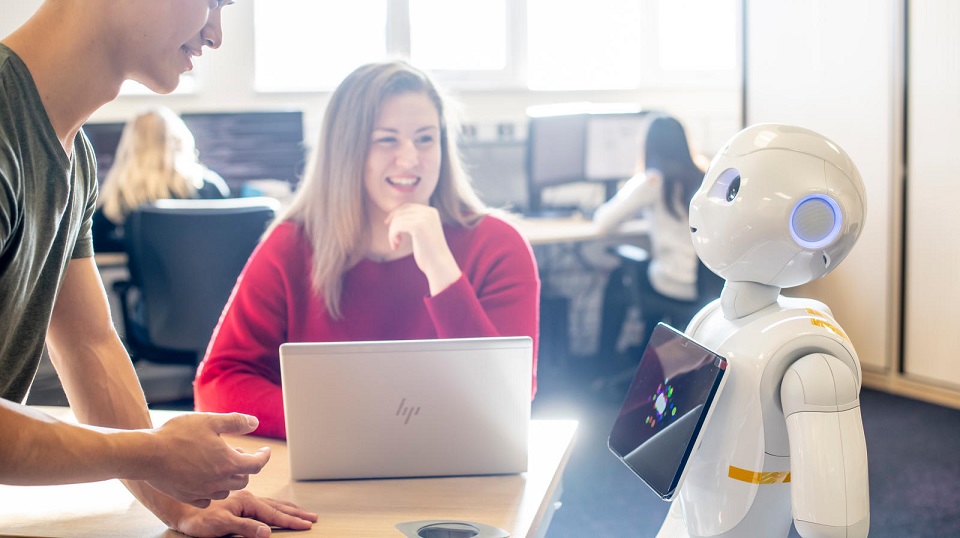 Female student Emma sat down at a table smiling at humanoid robot Pepper. Male student can be seen part in the image leaning on the table.