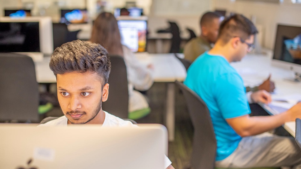 Student sat at a computer with several other students blurred in the background sat in the computer lab.